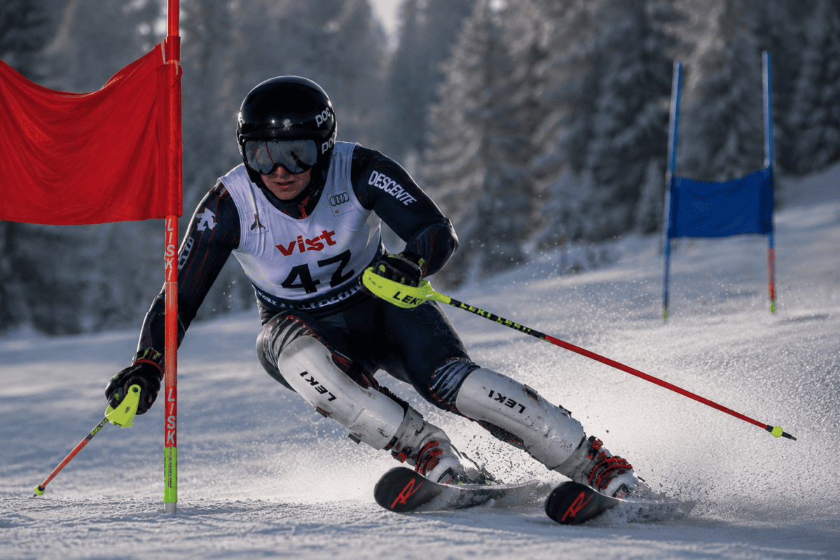 Alpine skier in white racing bib 47 carving a turn around a red slalom gate on a snowy course, goggles and helmet on the dark suit visible at speed within a pine forest.